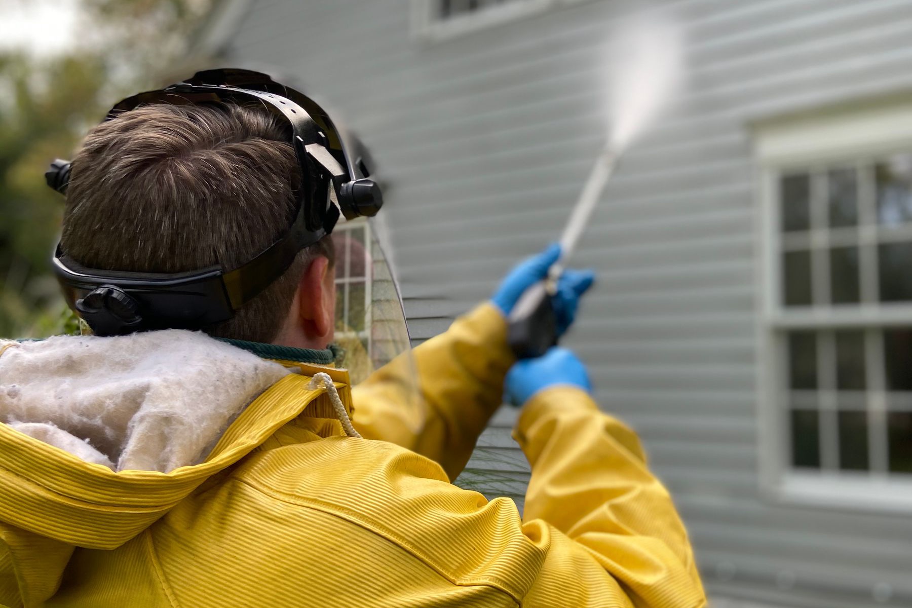 Person power washing house siding, seen from behind in a protective raincoat